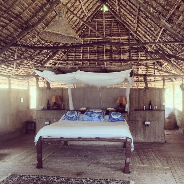 A rustic room with a thatched roof, a bed with a canopy, and a nightstand with a lamp. - Mikes Camp Kiwayu (Kiwayu)