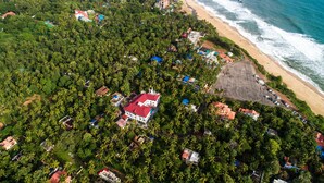An aerial view of a coastal area with dense greenery, a beach, and a large building with a red roof. - Sajjoys Hotel (Varkala)