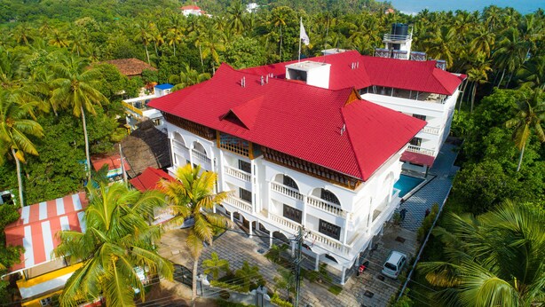 A multi-story building with a red roof, surrounded by lush greenery and palm trees. - Sajjoys Hotel (Varkala)