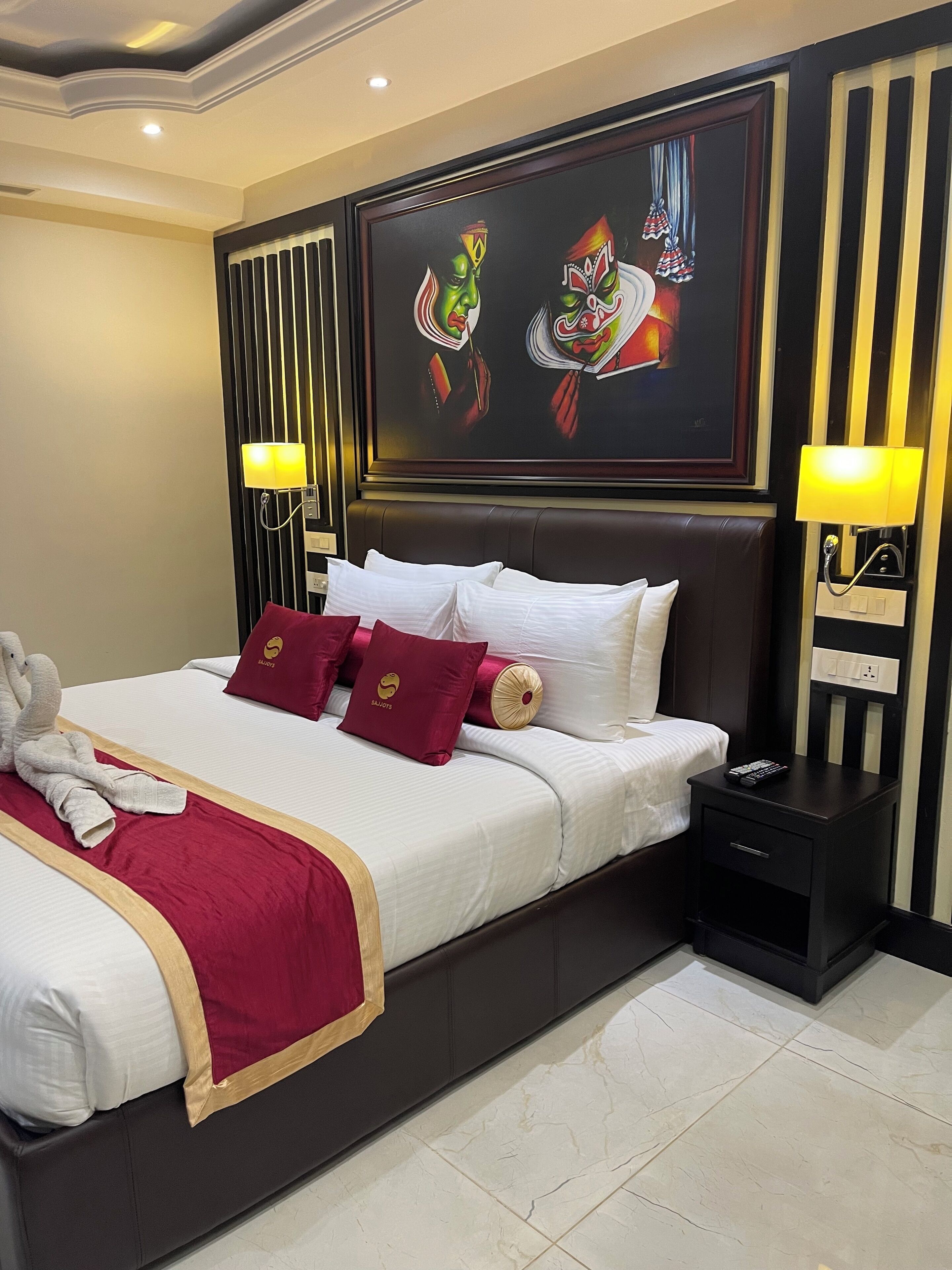 Ein Hotelzimmer mit einem großen Bett, weißer Bettwäsche, roten Kissen, einer roten Tagesdecke, einem Kopfteil mit einem Gemälde, zwei Nachttischlampen und einem Nachttisch mit Fernbedienung.