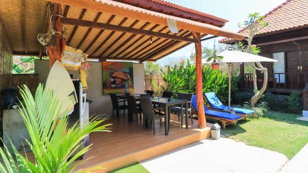Un patio couvert avec une table et des chaises en bois, un fauteuil bleu et un petit jardin agrémenté de plantes et d’un arbre.
