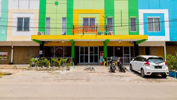 A multi-story building with a green and yellow facade, a covered entrance, and a parking area with a white car and motorcycles. - Botania Homestay (Batam)