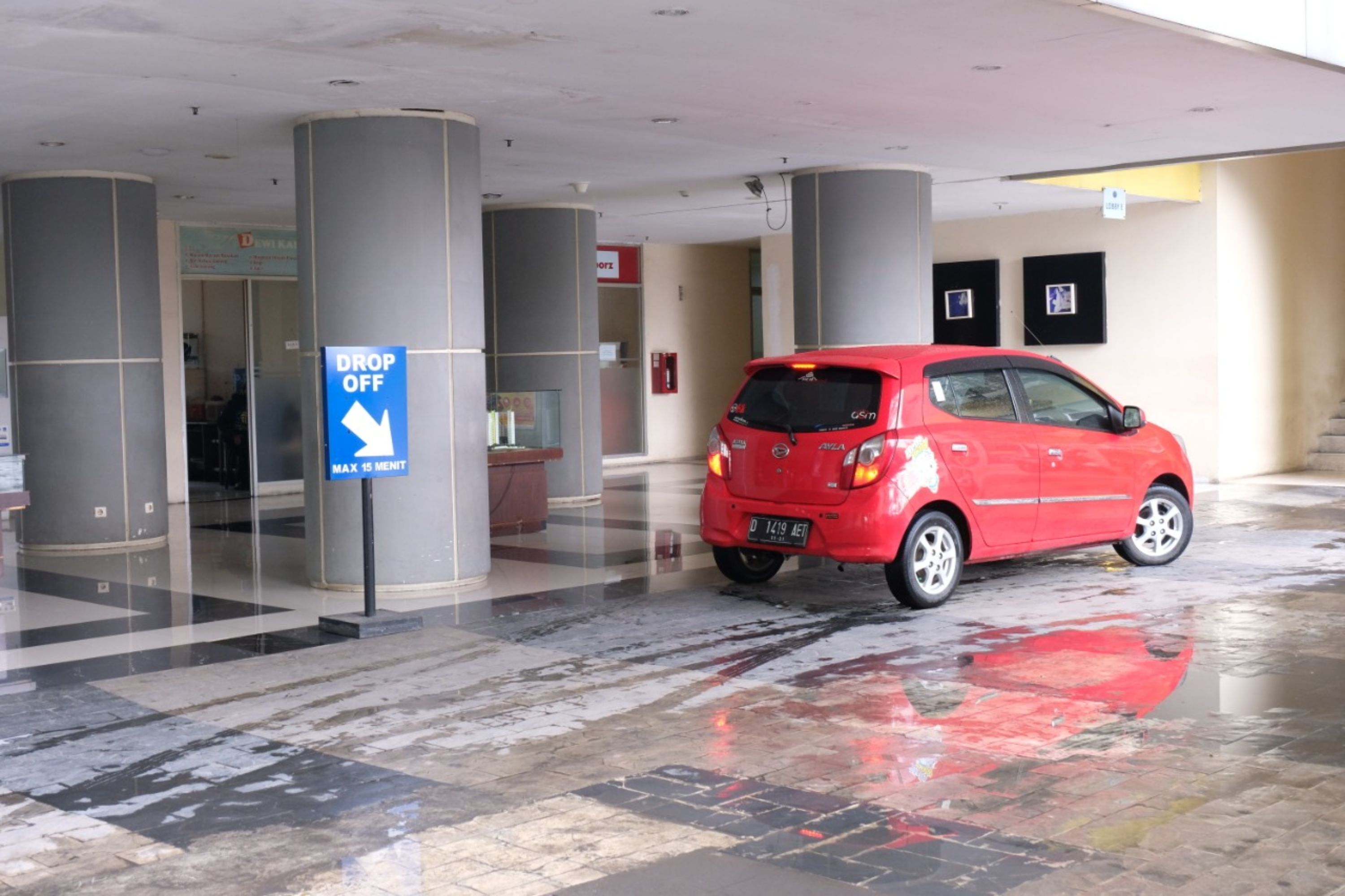 A red car parked in a covered parking area with a 'DROP OFF' sign and a 'MAX 15 MENT' sign.