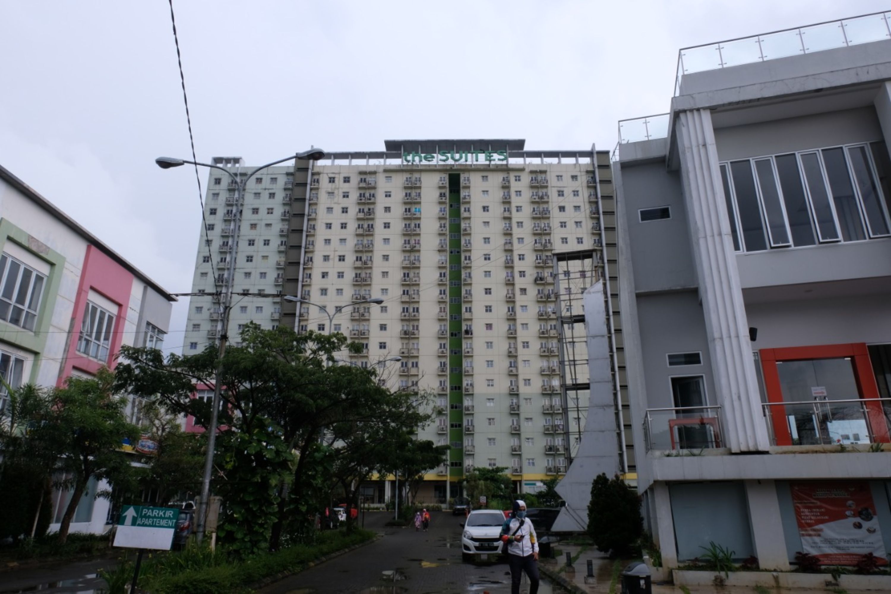 Un immeuble de grande hauteur, composé de plusieurs étages, avec une enseigne indiquant « PARKIR APARTMENT » au rez-de-chaussée, et un bâtiment moderne doté d’un balcon sur le côté droit.