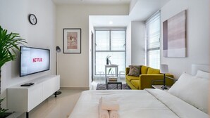 Een moderne hotelkamer met een bed, een televisie waarop 'NETFLIX' staat, een gele fauteuil en een groot raam met jaloezieën.