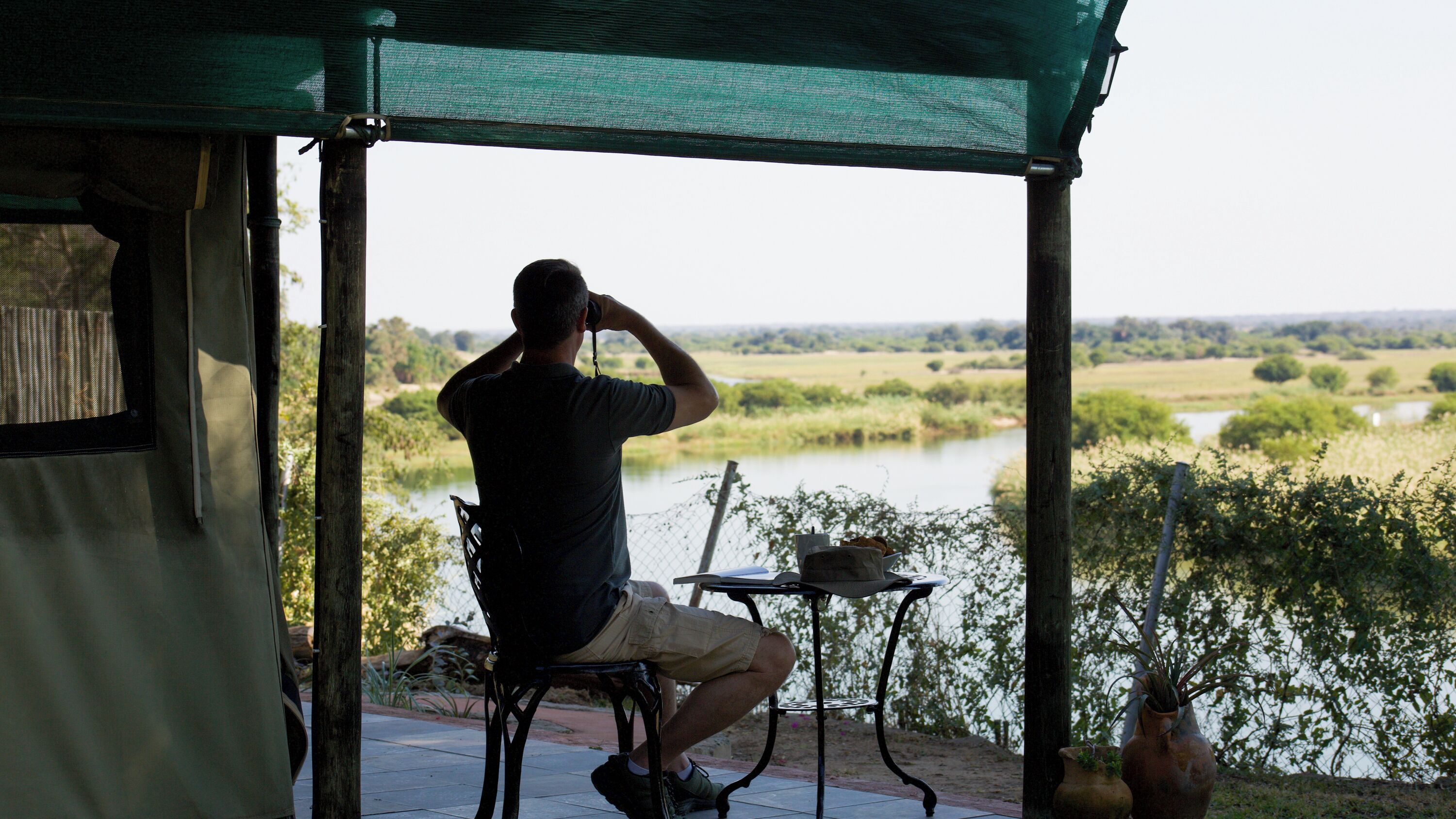 A man sitting at a table with a view of a river and fields.