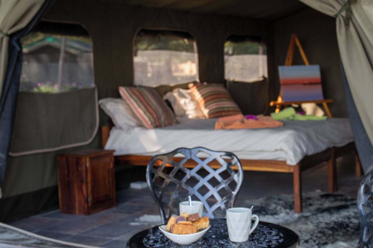 A tented accommodation with a bed, a table with a mug and cookies, and a painting on the wall.