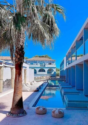 A modern pool area with a palm tree, a building with arched windows, and a clear blue sky.