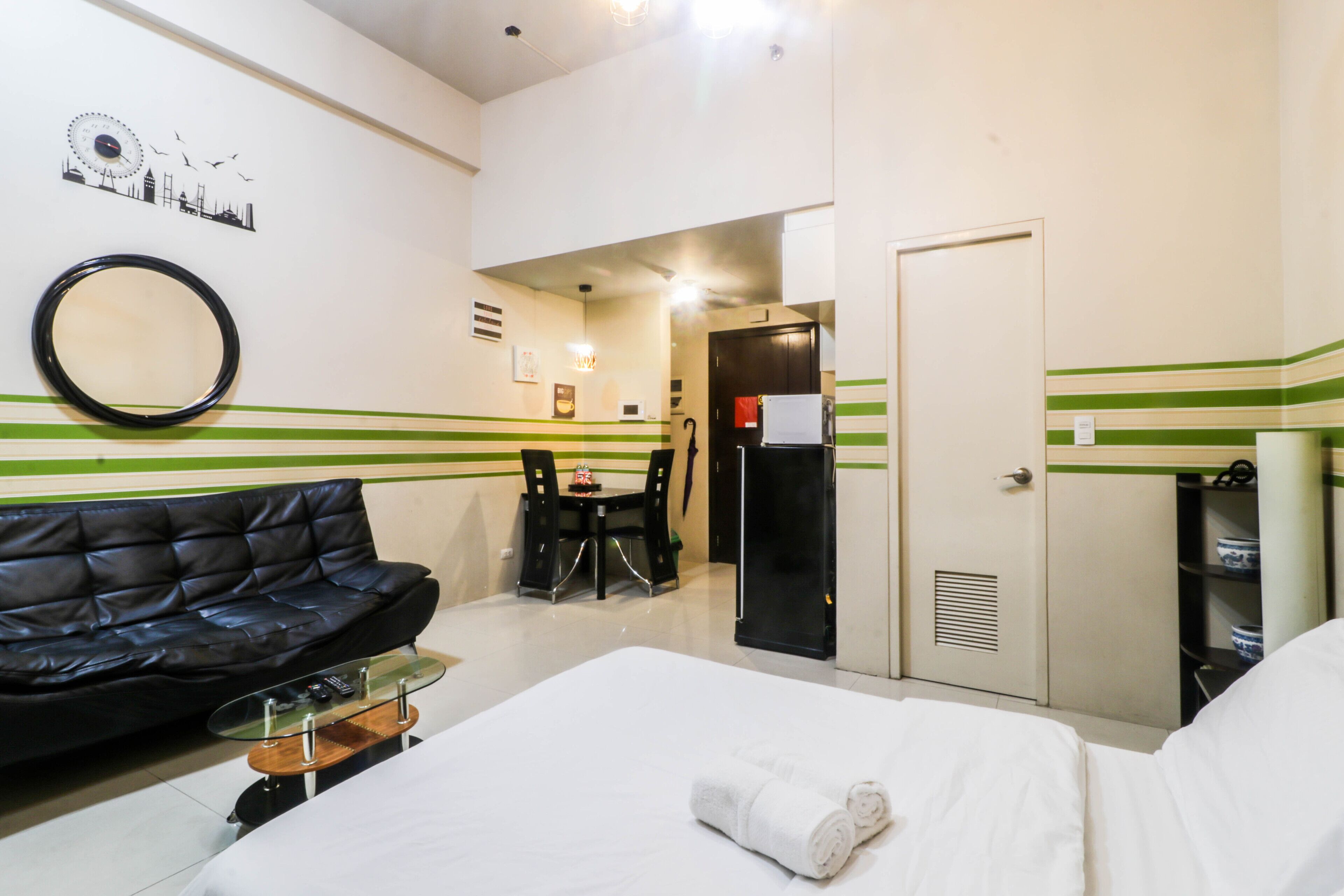 A hotel room with a bed, a black leather sofa, a small table, a mirror, a green-striped wall, a door, and a kitchenette area.