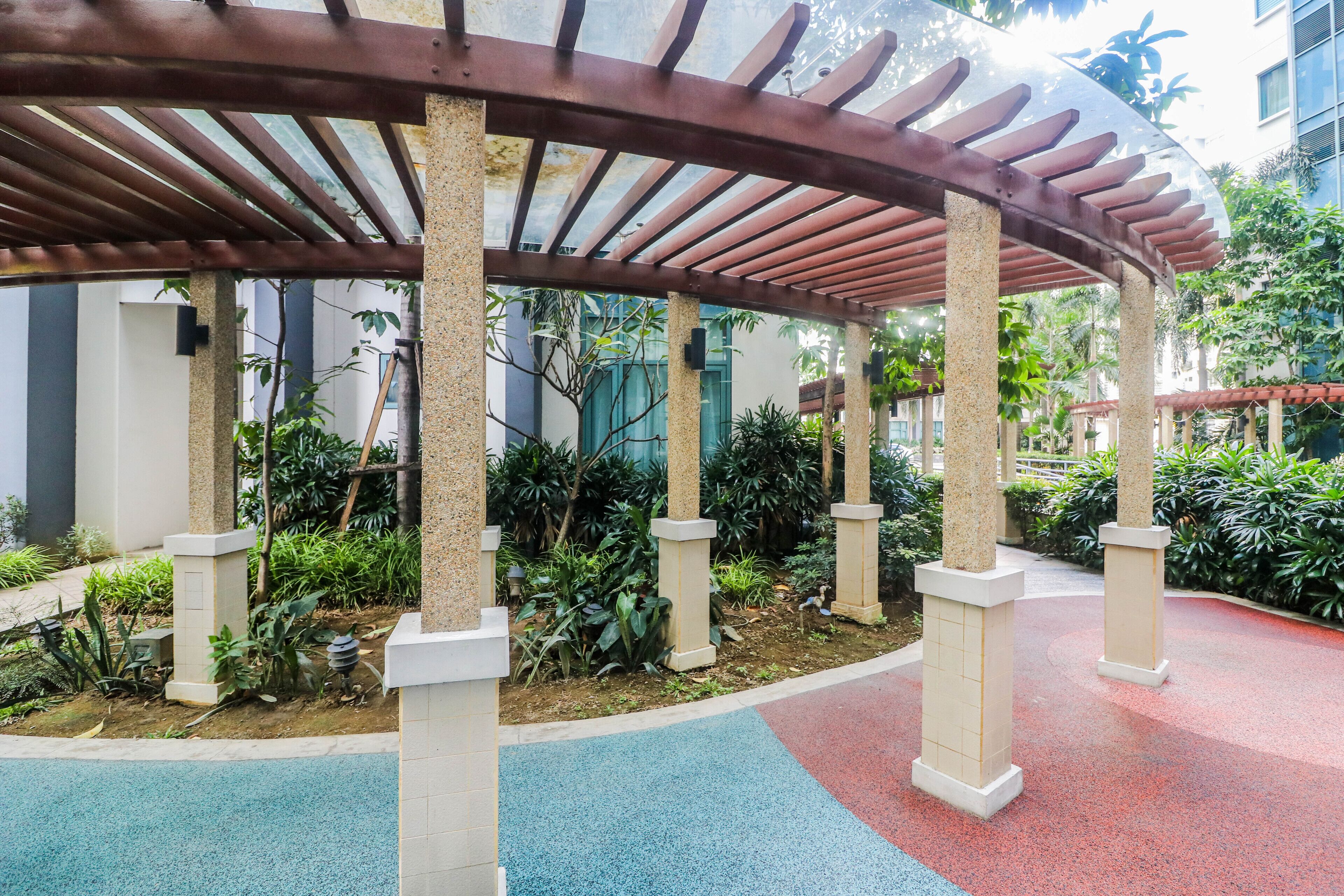 A covered outdoor area with a pergola structure, surrounded by greenery and a tiled floor.
