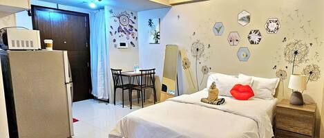 Habitación de hotel con cama, escritorio, silla, refrigerador y un muro decorado con piezas de arte hexagonales.