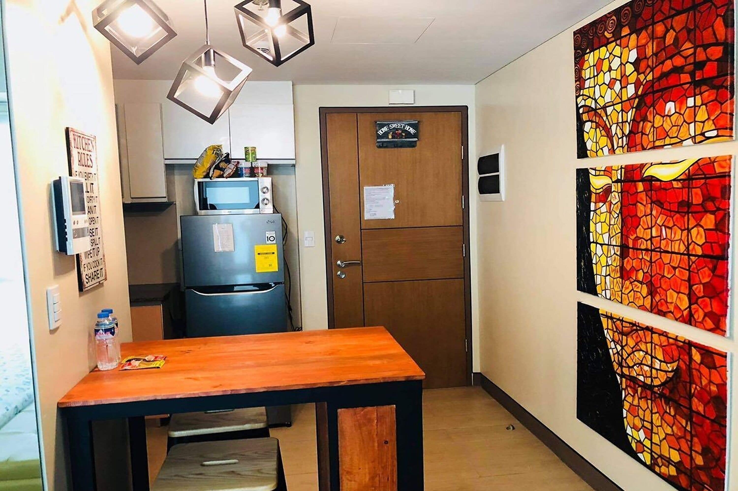 Eine kleine Küche mit Kühlschrank, Mikrowelle und einem Holztisch. Eine Tür mit einem Schild ist sichtbar, und ein farbenfrohes Mosaikkunstwerk befindet sich an der Wand.