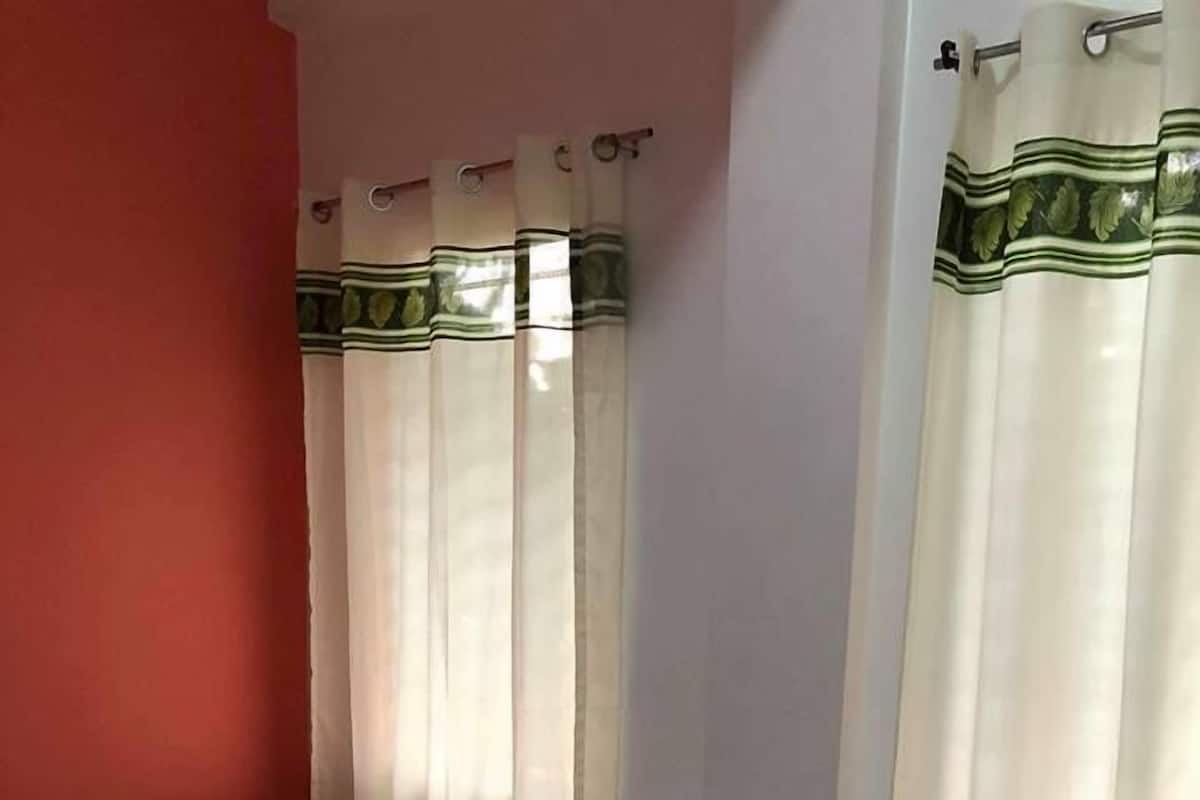 A single bed with a colorful pillow and a rolled towel, white curtains with green trim, and a red wall.