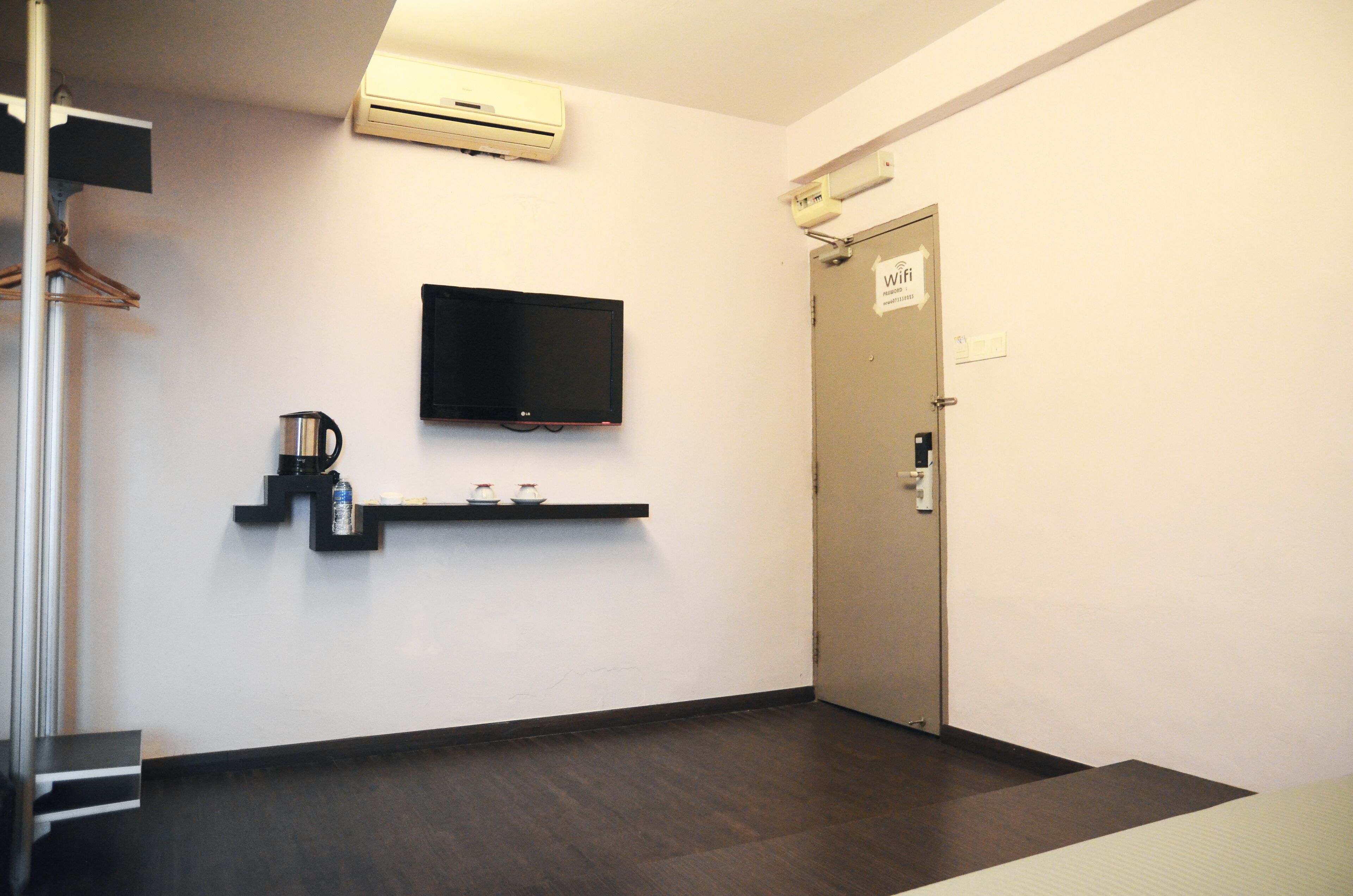 A hotel room with a flat-screen TV mounted on the wall, a coffee machine, and a shelf with a cup and a bottle.