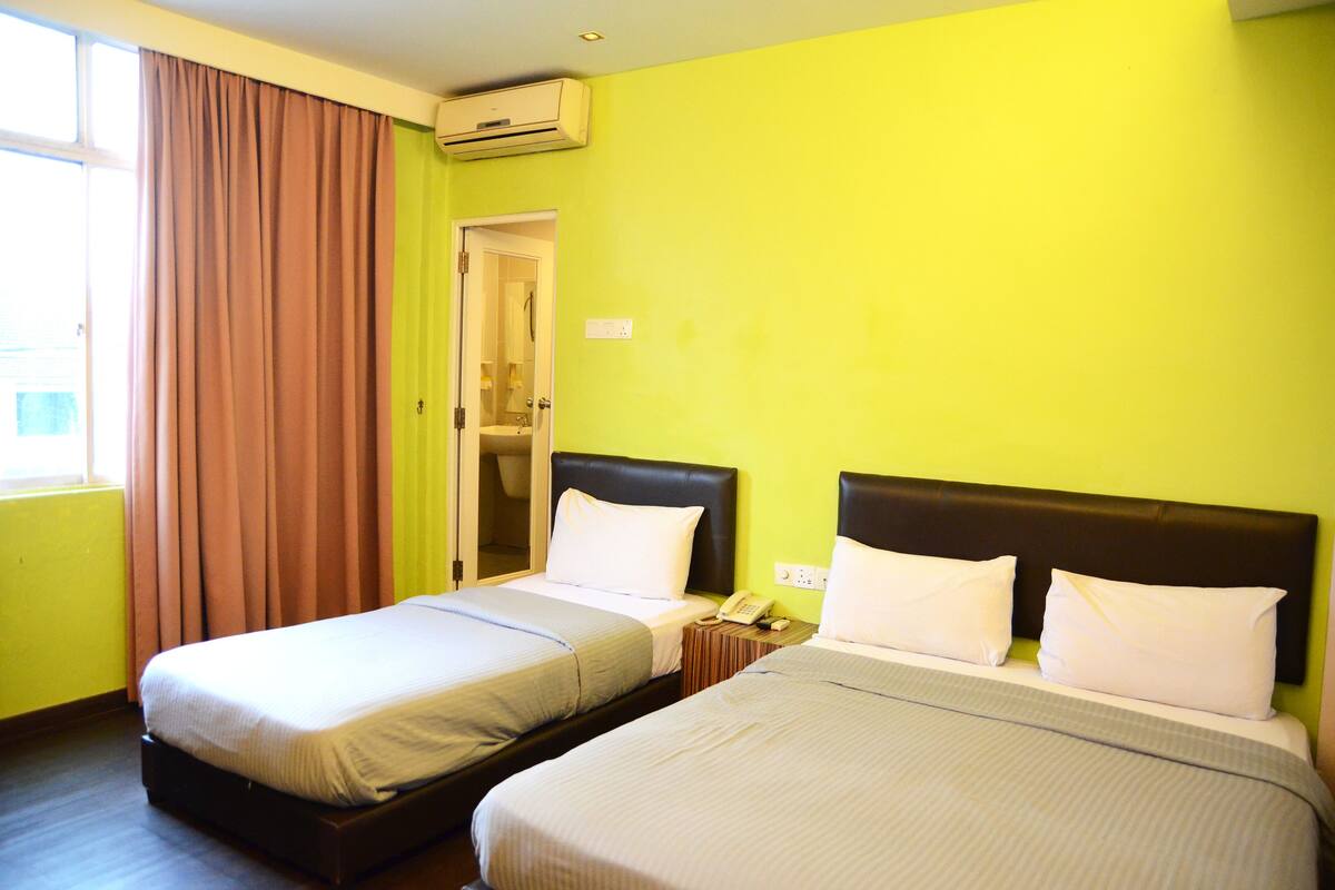 A hotel room with two single beds, a window with curtains, an air conditioning unit, and a door leading to a bathroom.