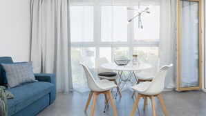 Uma sala de estar clara e moderna, com um sofá azul, cadeiras brancas, uma mesa de jantar redonda e janelas amplas com cortinas brancas.