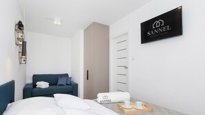 Quarto de hotel moderno com cama, sofá azul, televisão branca montada na parede exibindo o logotipo SANNEL e cama bem arrumada com bandeja de itens de café da manhã.