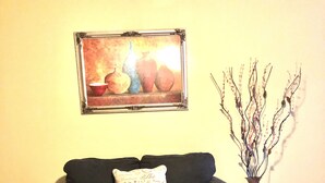 Una sala de estar con un sofá de color oscuro, una mesa de centro redonda de madera y un jarrón con flores blancas, además de un cuadro en la pared.