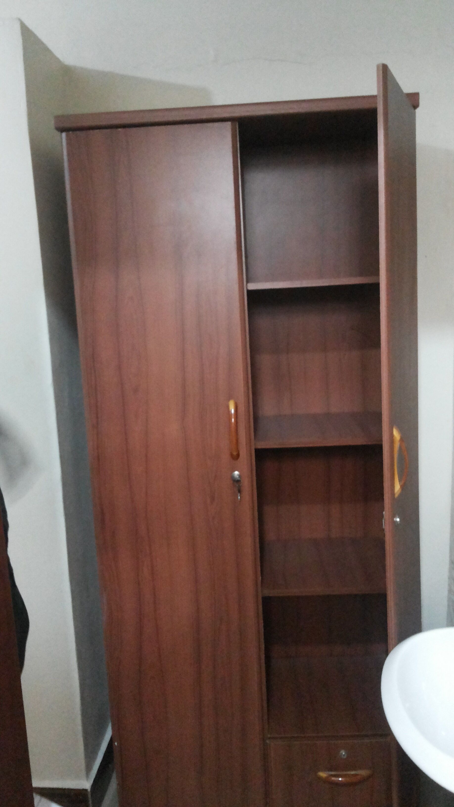 Une armoire en bois, dotée de plusieurs étagères et d’un seul tiroir, se trouve dans une pièce où se trouve un lavabo blanc.