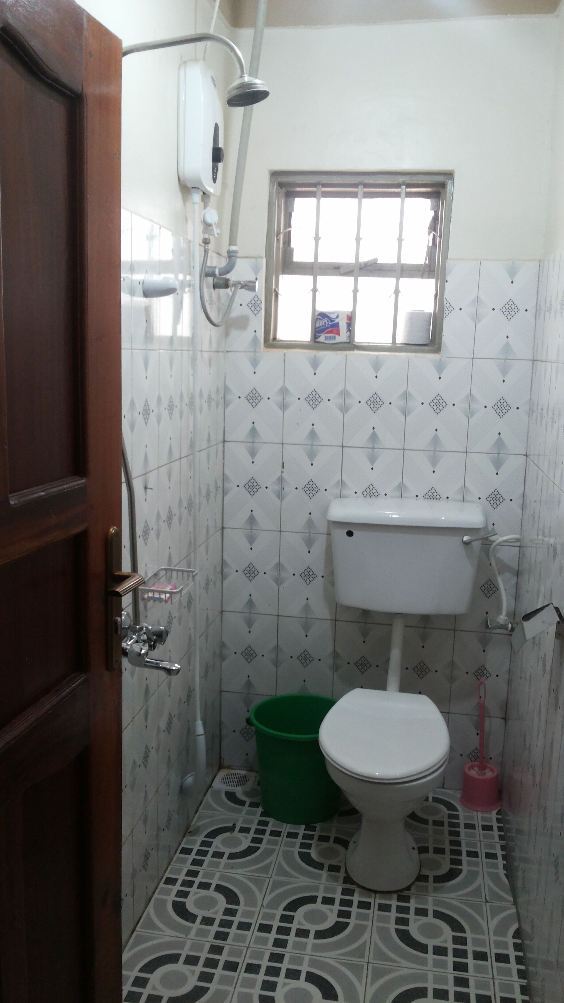 A small bathroom with a toilet, a green trash bin, a showerhead, and a window.