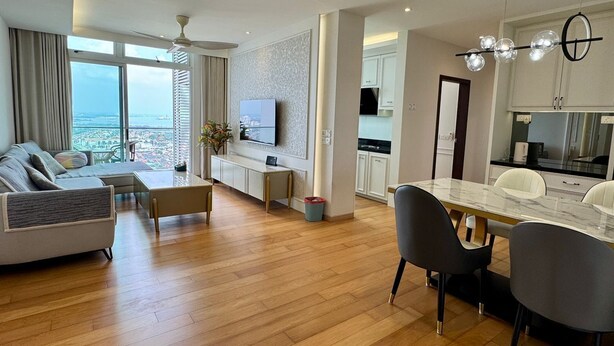 A modern living room with a sofa, dining table, and chairs, a TV, and a view of the ocean. - JB City Shopping Mall Apartment (Johor Bahru)