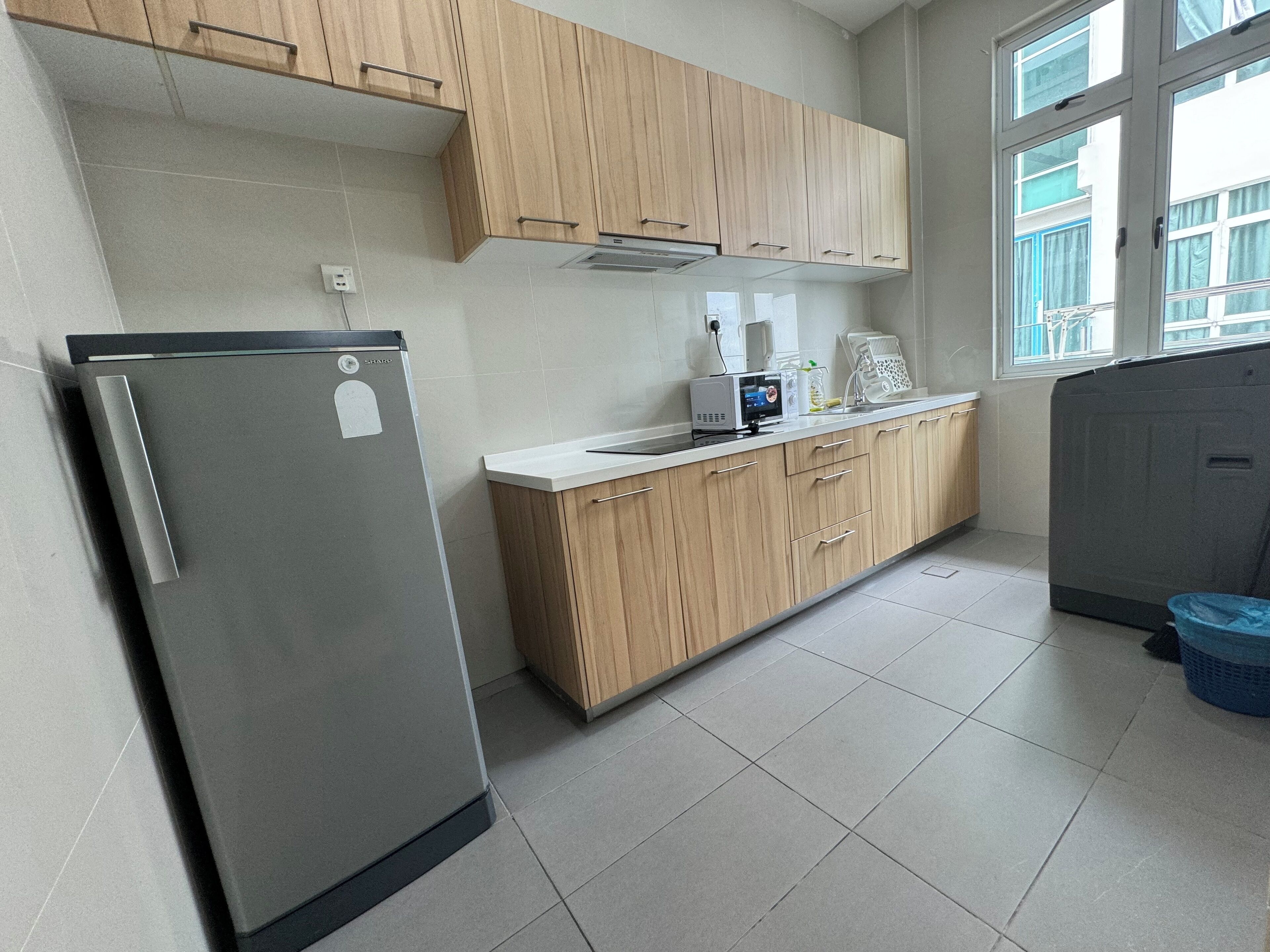 A kitchen with wooden cabinets, a stainless steel refrigerator, a microwave, and a window with a view of buildings outside.