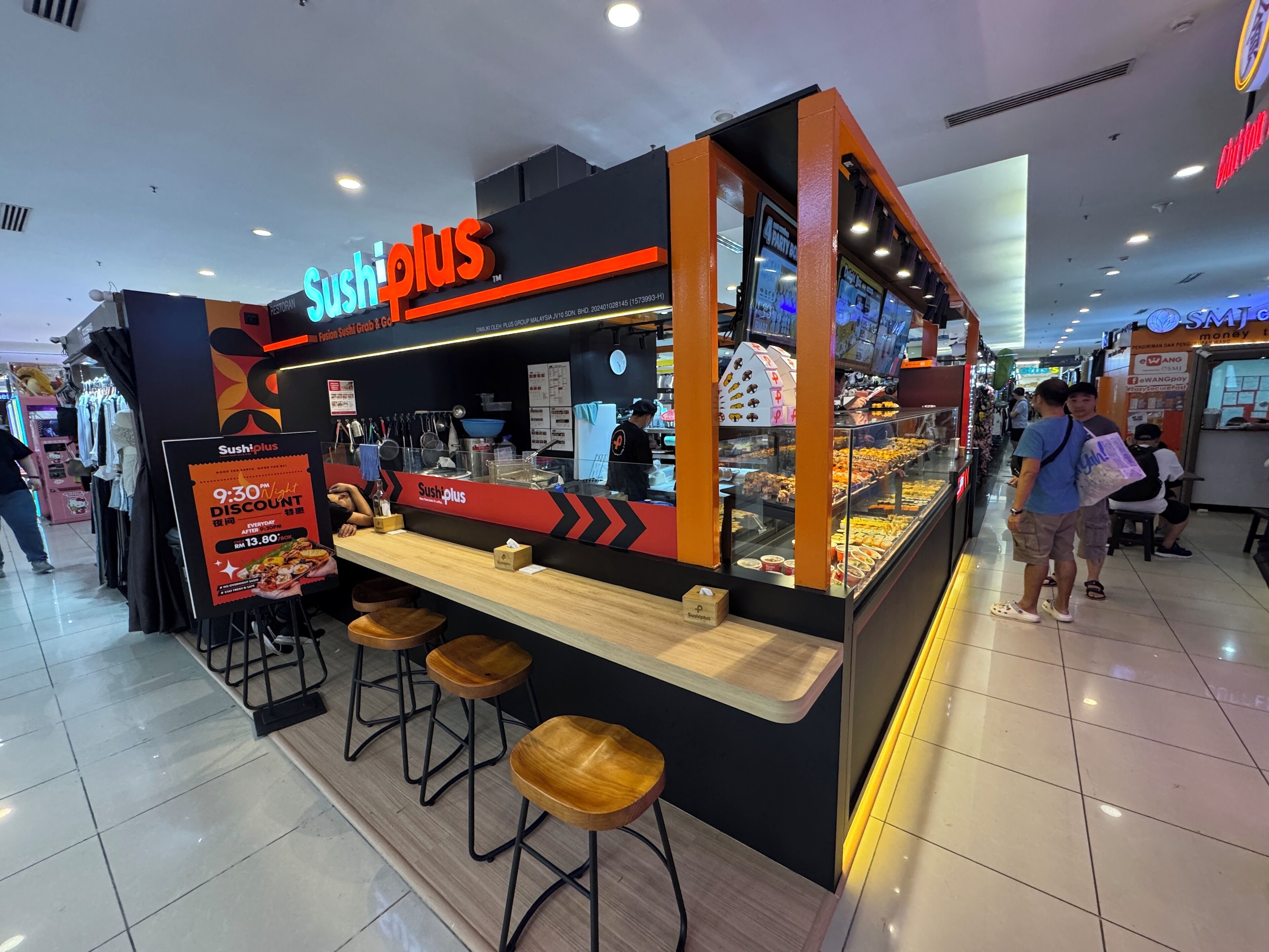 A sushi bar named 'Sushi Plus' with a glass display of various dishes, a promotional sign offering a discount, and wooden stools.
