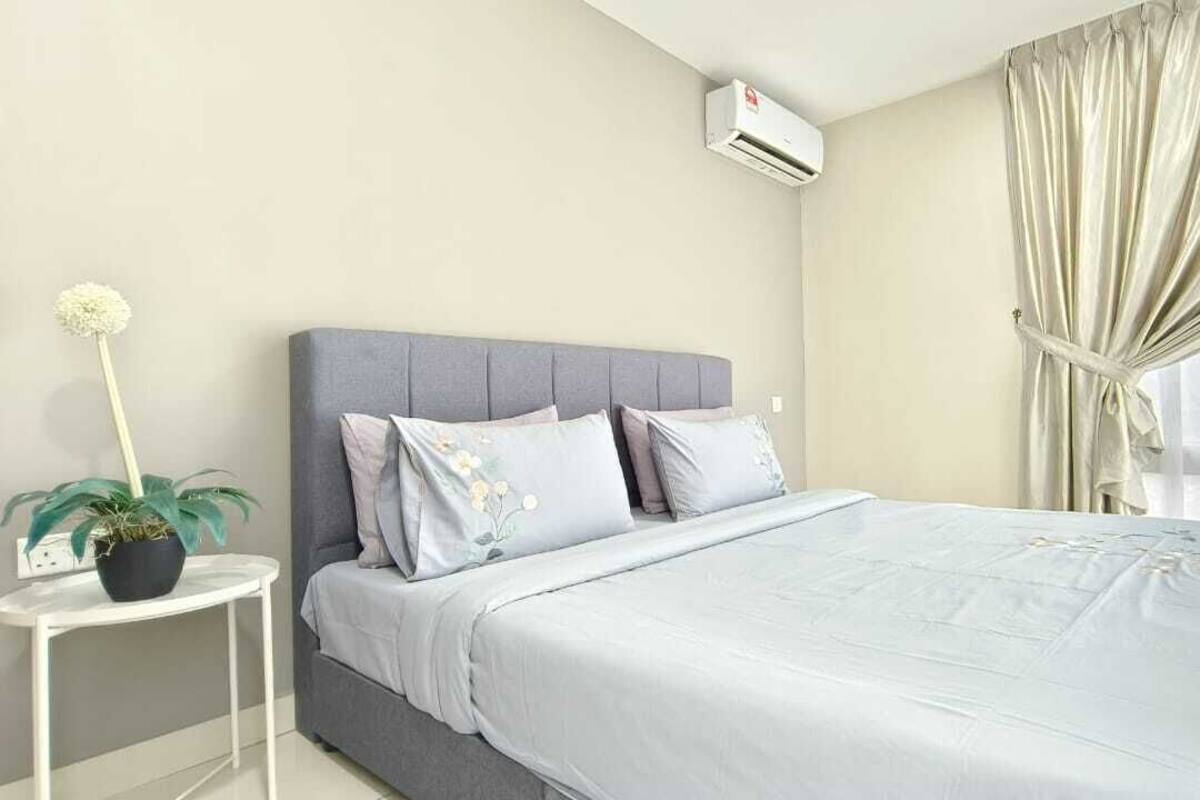A bedroom with a grey upholstered headboard, a bed with a light blue bedspread, a small white bedside table with a potted plant, and a window with sheer curtains.