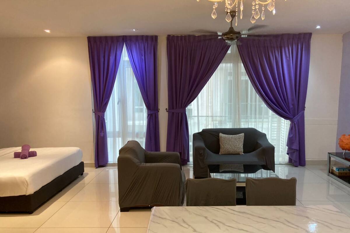 A modern hotel room with a bed, two armchairs, a sofa, a glass coffee table, and purple curtains.