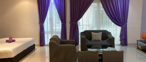 A modern hotel room with a bed, two armchairs, a sofa, a glass coffee table, and purple curtains.