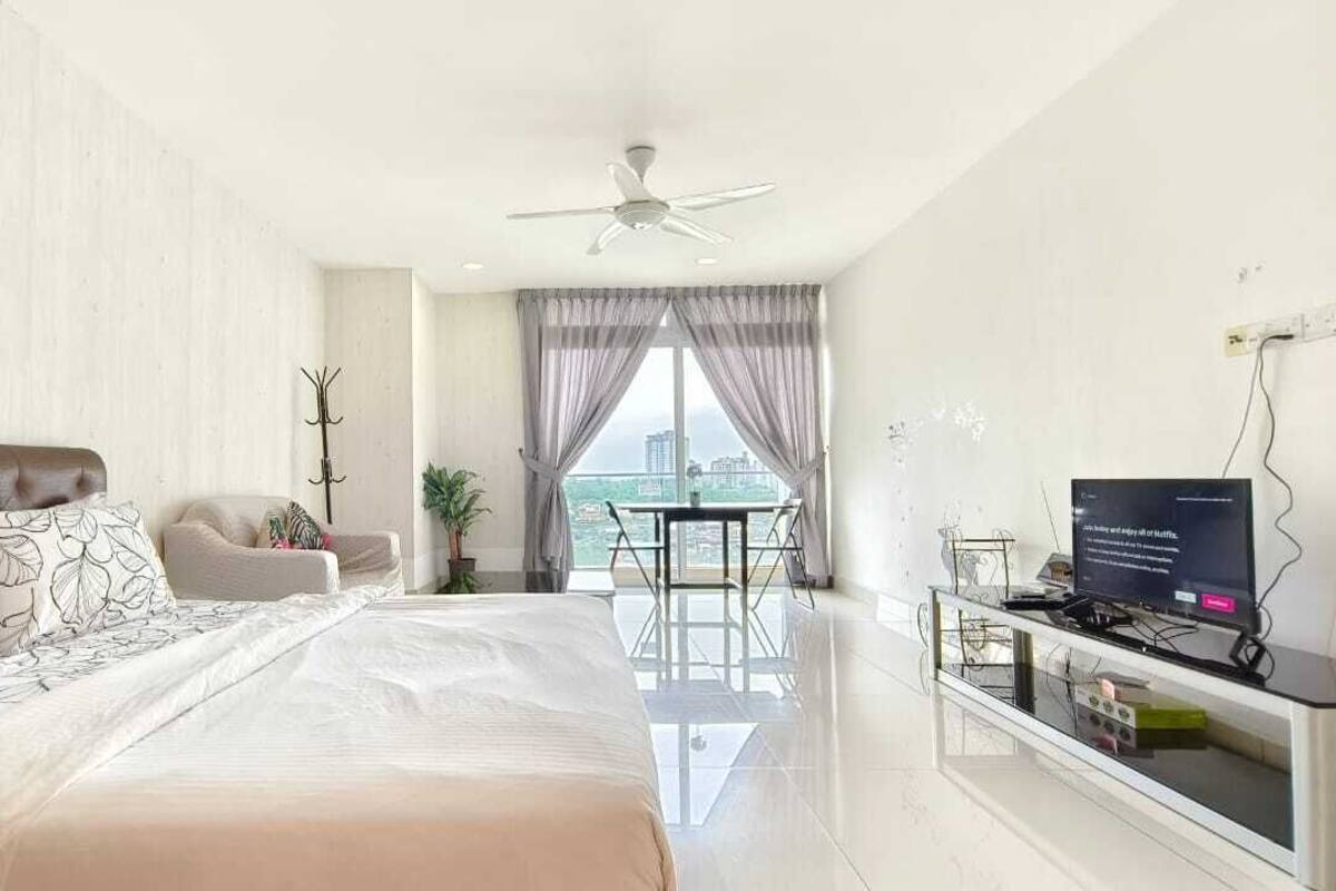 A bedroom with a large bed, a TV on a stand, a ceiling fan, and a view of the city through the window.