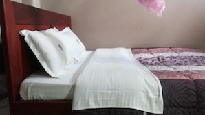 A bed with white bedding and a wooden headboard, a patterned blanket, and a pink decorative item. - GAF Apartments (Nebbi)