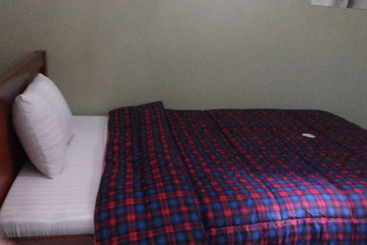 A bed with a plaid bedspread and a white pillow in a room with a plain wall and a wooden headboard.