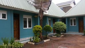 A building with a blue exterior, white windows, and a tiled roof. There are three small trees in front of the building. - GAF Apartments (Nebbi)