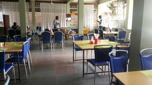 A dining area with tables and chairs, a person standing near the window, and another person walking in the background. - GAF Apartments (Nebbi)