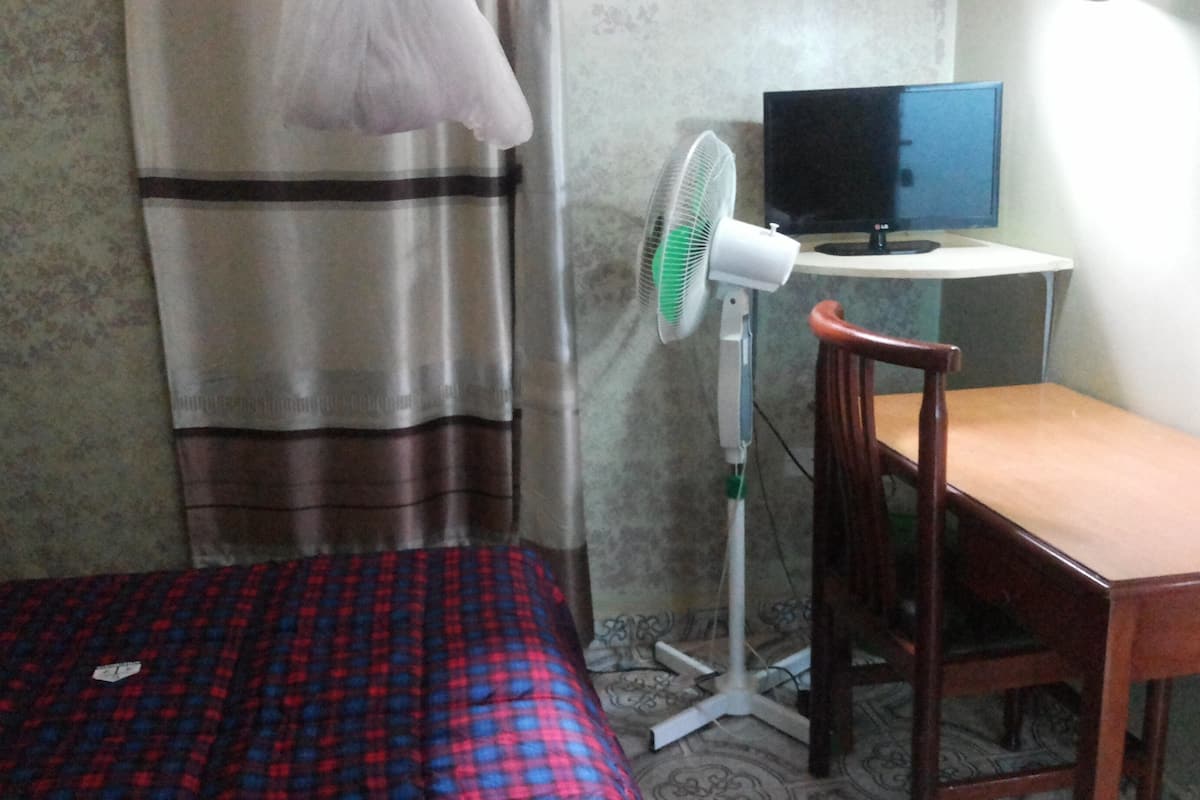 A room with a bed, a desk, a chair, a television, and a standing fan.
