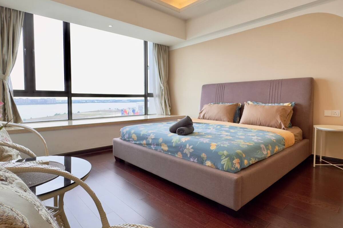 Ein Schlafzimmer mit einem großen Bett, Meerblick durch ein Fenster und ein Korbsessel.