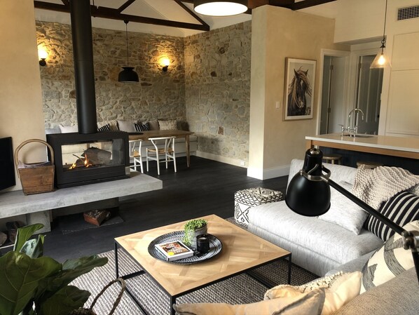 A cozy living room with a stone wall, a fireplace, a dining area, and a kitchen with a sink. - Belle’s Cottage- Luxurious, Stirling escape with cosy fire, deck, tennis court  (Aldgate)