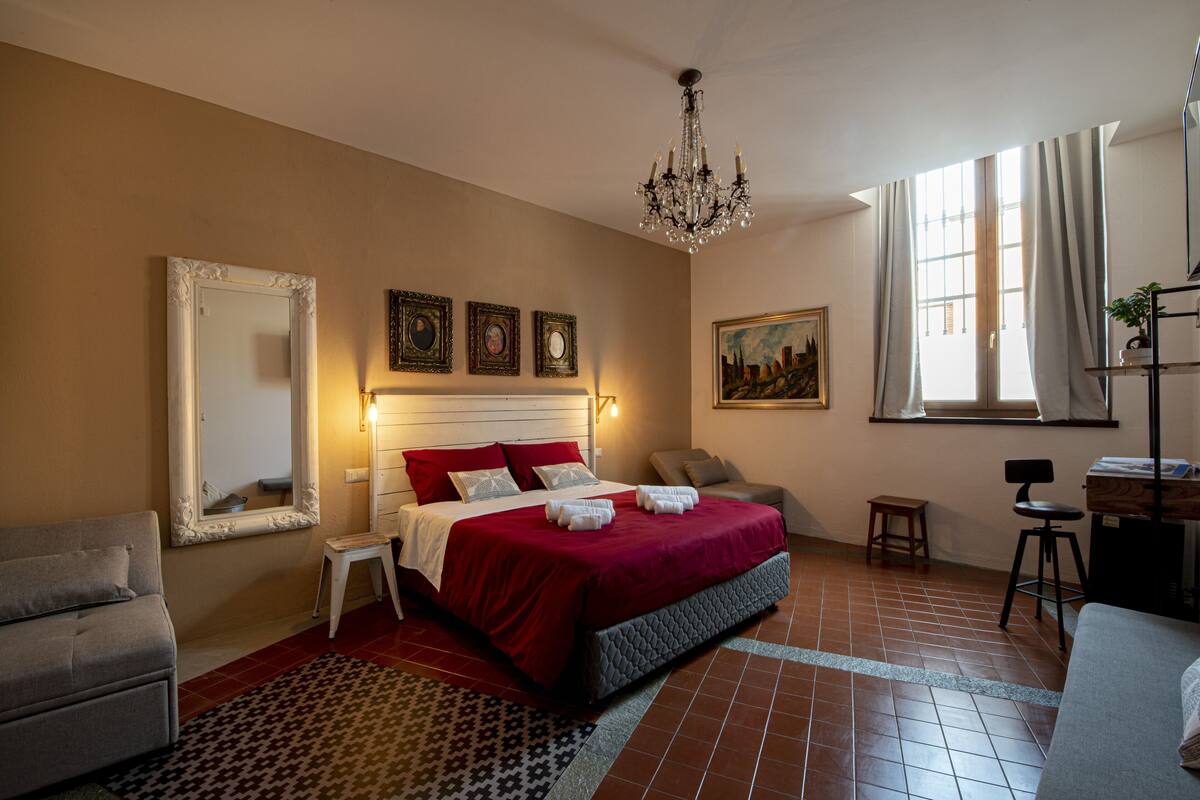 A bedroom with a large bed, a chandelier, a sofa, a chair, a small table, and a painting on the wall.