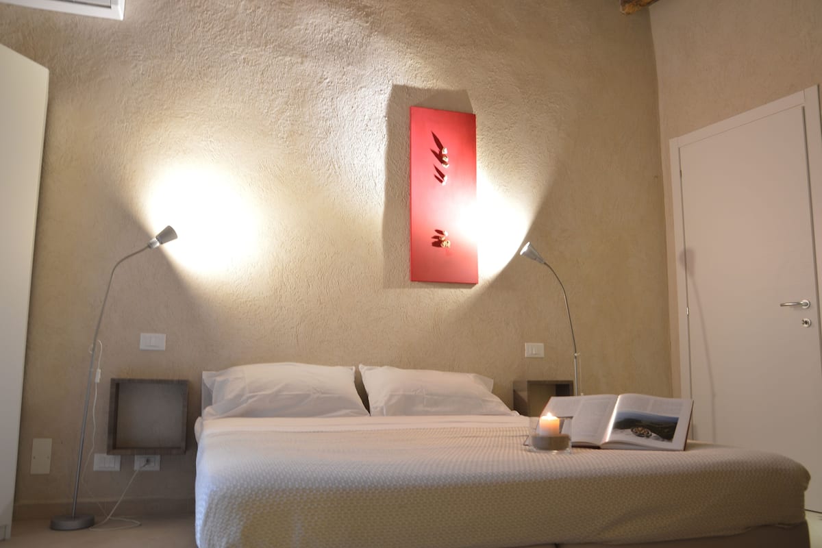 A neatly made bed with white linens, a bedside lamp, a book, and a small candle on the nightstand.