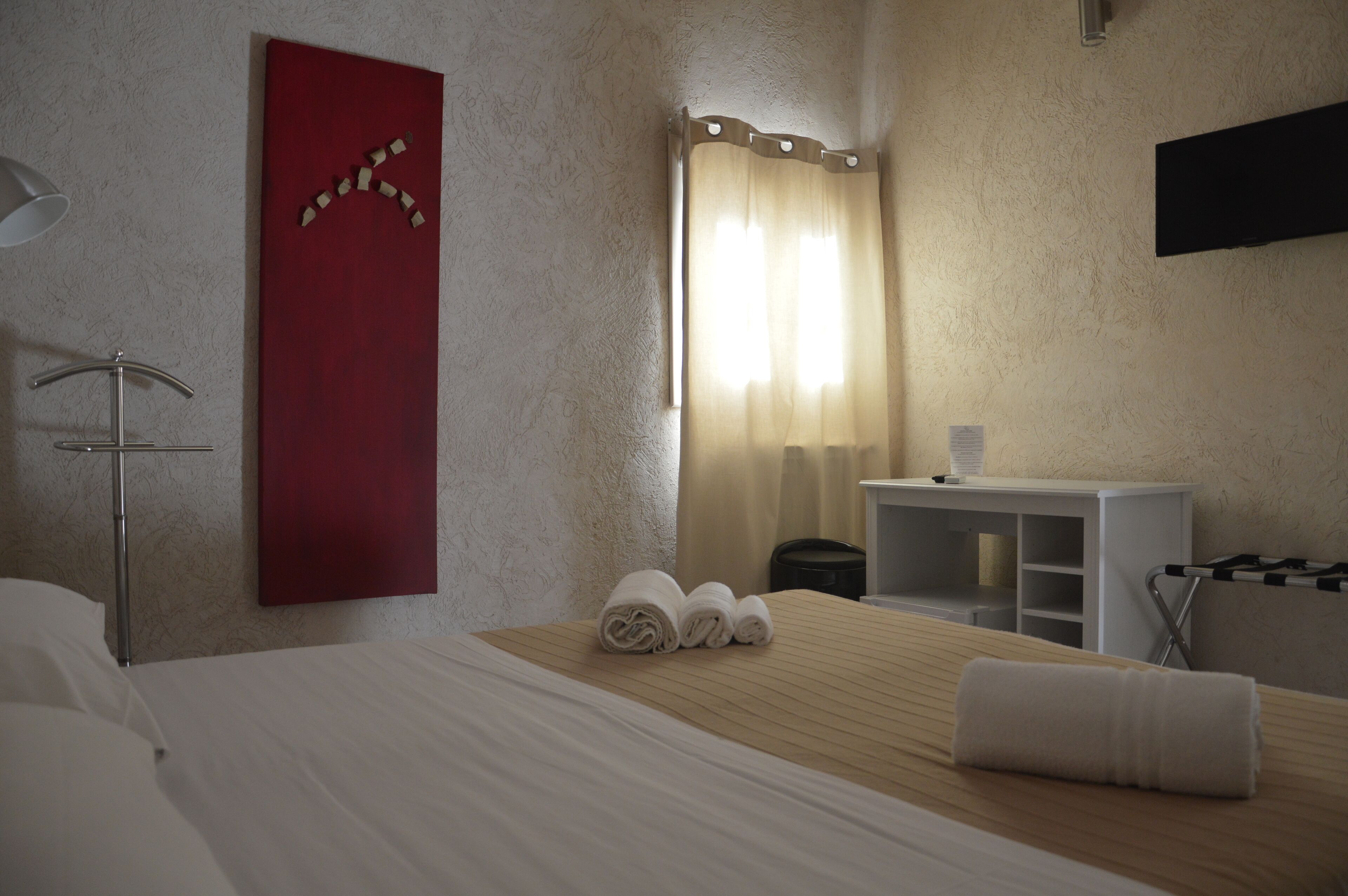A hotel room with a bed, a red wall decoration, a window with curtains, a desk, and a TV mounted on the wall.