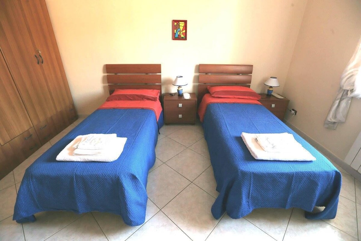 A room with two beds, each with a blue bedspread and red pillows, and a wooden wardrobe.