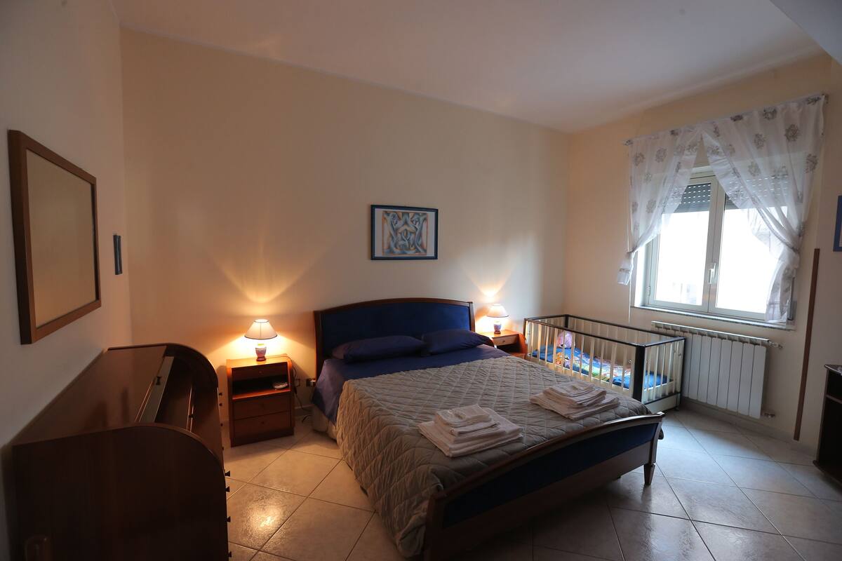 A bedroom with a bed, bedside lamps, a crib, a radiator, and a window with curtains.