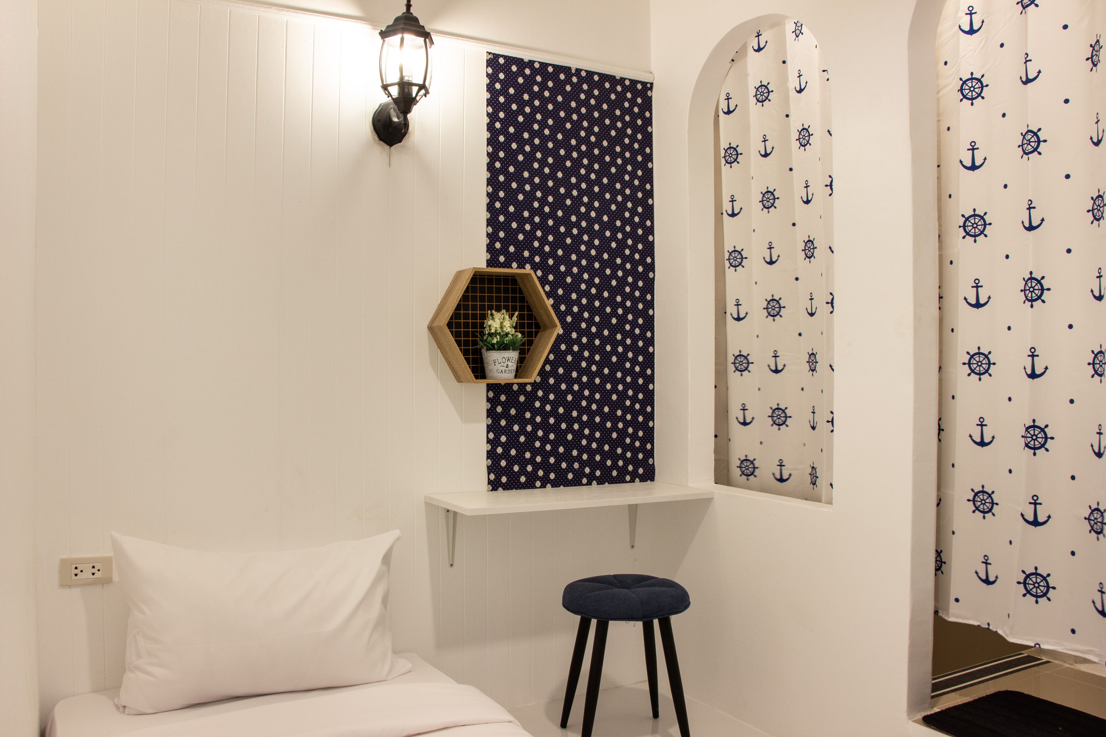 A bedroom with a bed, a nightstand, a wall-mounted shelf with a plant, a window with curtains, and a wall with a nautical pattern.