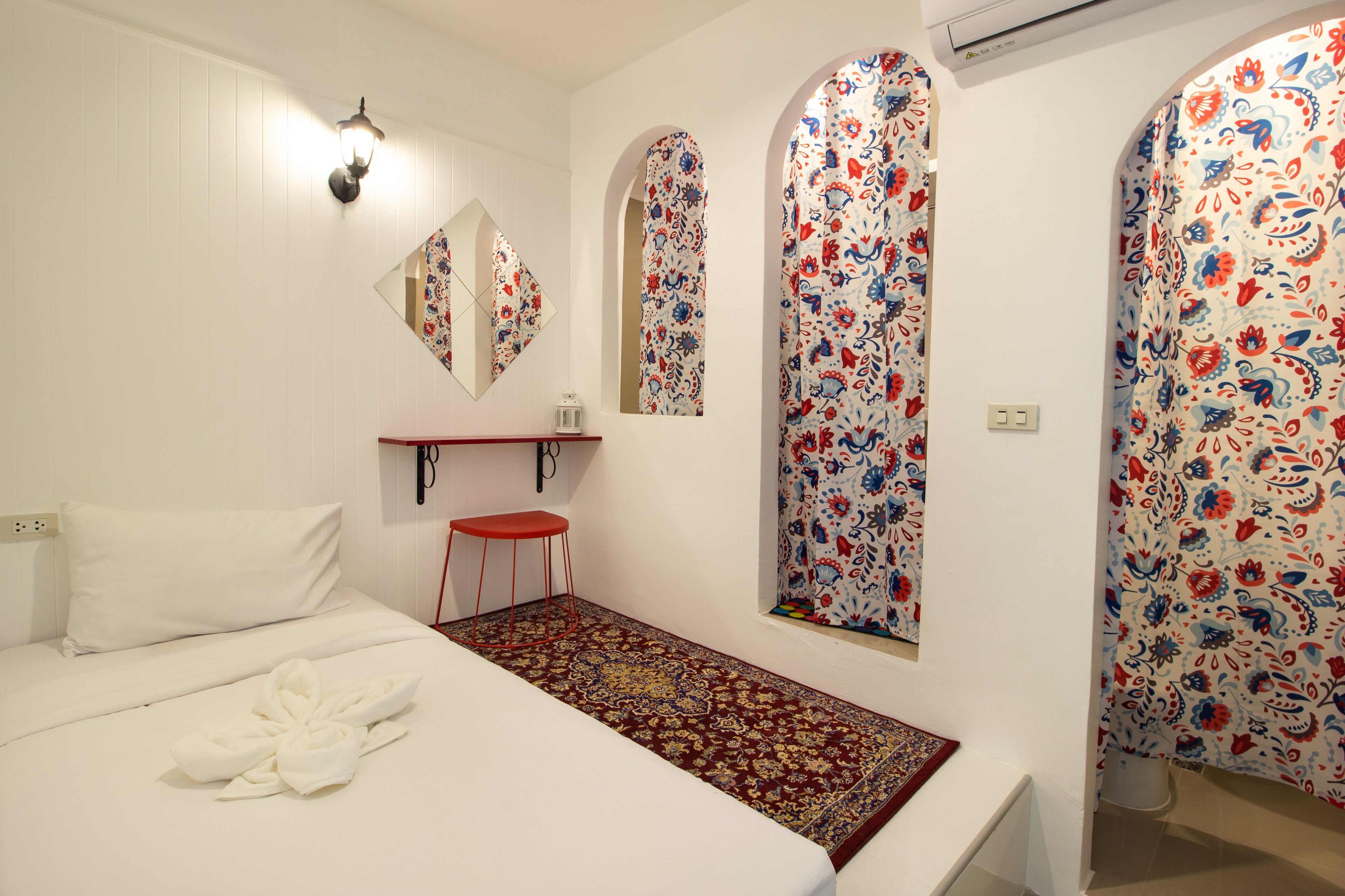 A hotel room with a bed, a red stool, a small table, a mirror, and a patterned window.