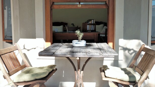 Une table en bois sur laquelle est posée une plante en pot, deux chaises en bois avec des coussins, et une vue sur une pièce comprenant un bureau et des livres.