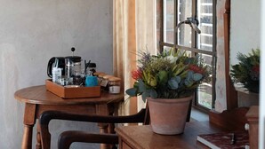 Une table en bois, sur laquelle se trouvent une machine à café, des bocaux et une boîte de café, est placée près d’une fenêtre ornée de rideaux et d’une plante en pot.