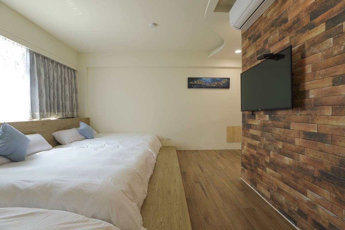 A bedroom with a bed, a flat-screen TV mounted on a brick-patterned wall, and a window with curtains.