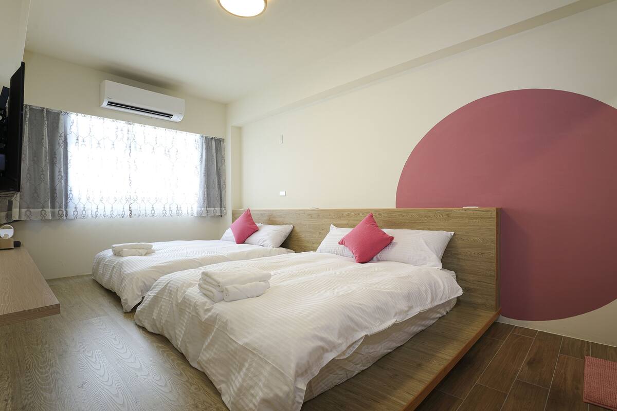 A hotel room with two beds, a wooden headboard, a large window with curtains, an air conditioning unit, and a wall with a large red circle.