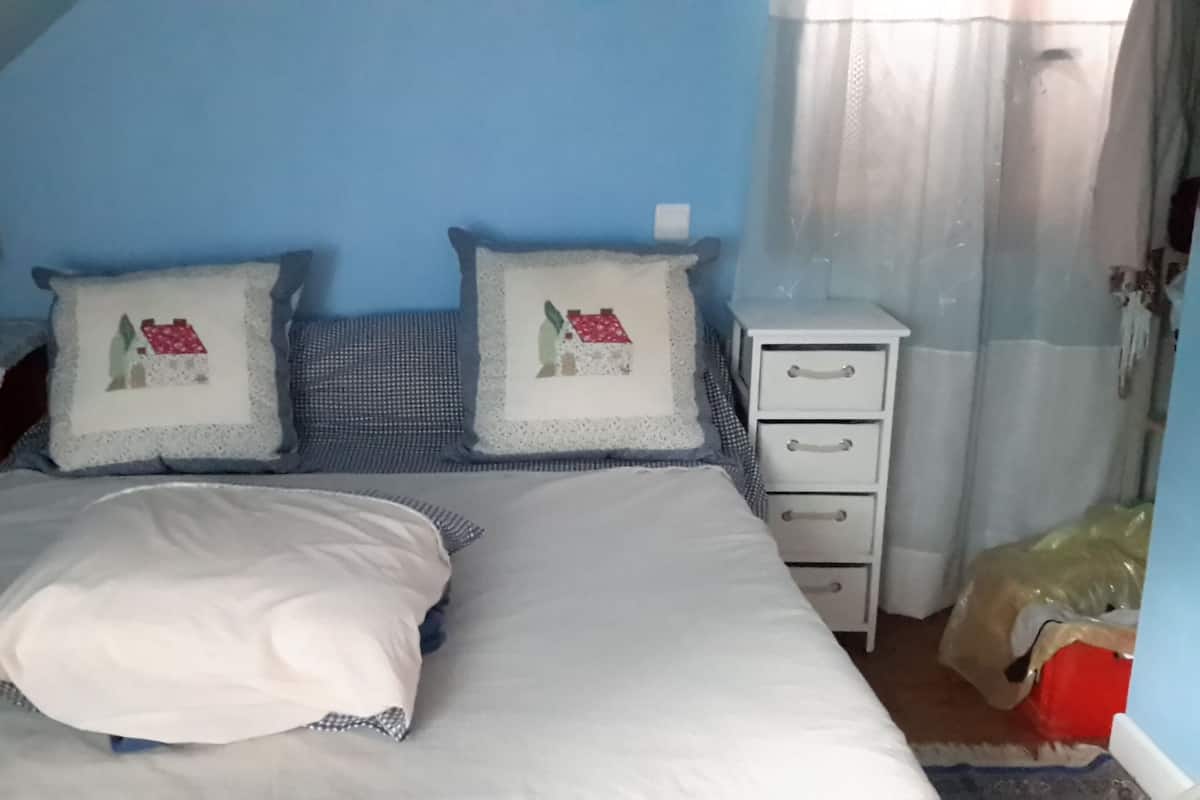 Een slaapkamer met een bed, twee kussens, een nachtkastje, een lamp en een raam met gordijnen.